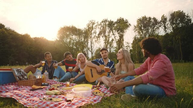 Group of multi-ethnic teenagers toast and celebrate during a picnic in the park. Millennials sing songs while playing the guitar and share a positive mood.