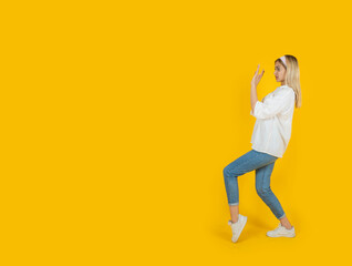Fototapeta na wymiar Doing stop gesture, side full length body view 20s blonde caucasian girl doing stop gesture. Showing open palm aside copy space. Personal space sign, say no concept idea image. Yellow background.