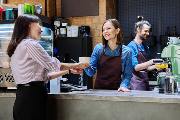 Woman customer of coffee shop near counter with cup of coffee talking to restaurant workers