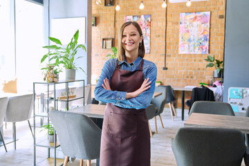 Young woman worker, owner in apron looking at camera in restaurant, coffee shop