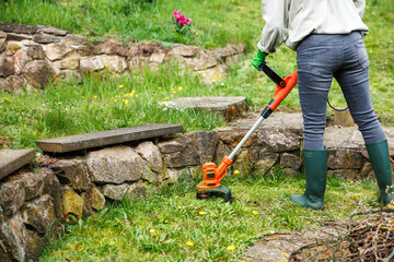 Woman is trimming grass next stone wall in garden. Lawn care by grass trimmer