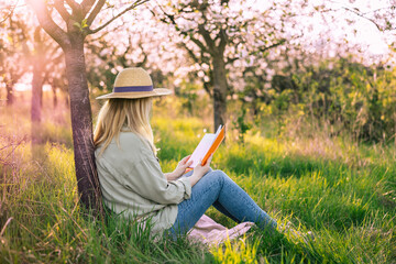 Relaxed woman sitting under blooming cherry tree and reading book to improve her mindfulness. Enjoying moments of solitude and relaxation in spring orchard