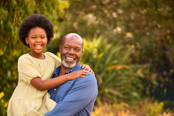 Portrait Of Loving Grandfather Laughing And Hugging Granddaughter Outdoors In Countryside Or Garden