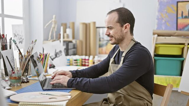 A focused man with a beard and ear piercing works in an art studio, using a laptop surrounded by brushes and paint.