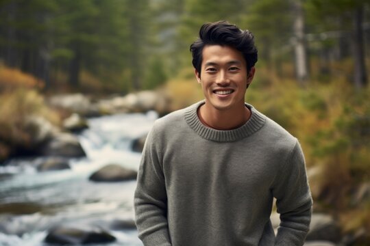 Portrait of a grinning asian man in his 20s dressed in a warm wool sweater on tranquil forest stream