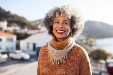 Portrait of a glad afro-american woman in her 50s dressed in a warm wool sweater while standing against picturesque seaside village - obrazy, fototapety, plakaty