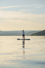 A man is standing and paddling on a paddleboard or SUP on the lake at sunset. Enjoying the vacation. Landscape in the background. Training SUP board.