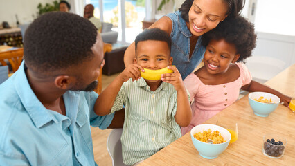 Son Having Fun Playing With Banana As Family Sit At Kitchen Counter Eating Breakfast At Home