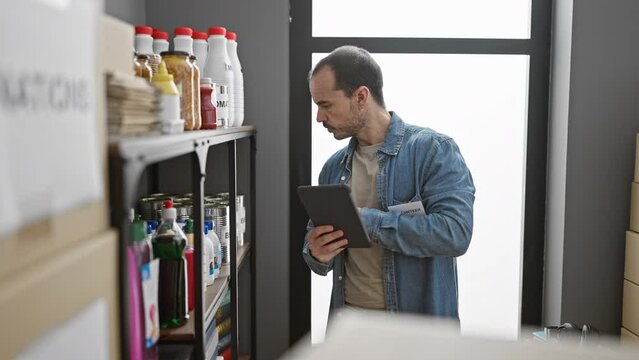 A bald man with a beard wearing a denim shirt uses a tablet in a storeroom with shelves stocked with food and cleaning supplies.