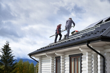 Fototapeta na wymiar Electricians installing solar panel system on roof of house. Men workers in helmets carrying photovoltaic solar module outdoors. Concept of alternative and renewable energy.
