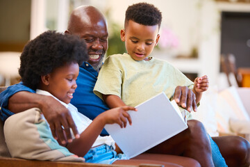Grandfather And Grandchildren Sitting On Sofa At Home Reading Book Together
