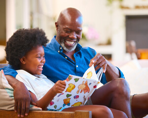 Family With Grandfather And Granddaughter Sitting On Sofa At Home Reading Book Together