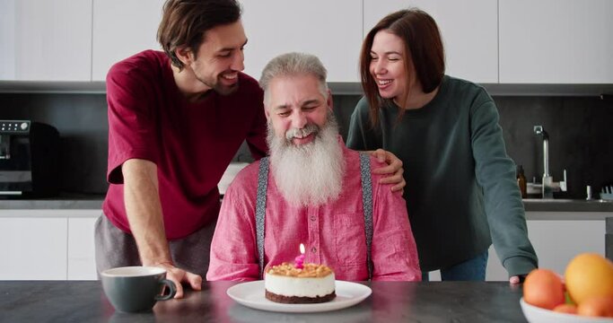 A happy elderly man with gray hair and a lush beard in a pink shirt blows out a candle on a cake and his adult children a man and a brunette girl in a green jacket congratulate their dad on his
