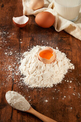 Preparation for healthy meal with a fresh egg cracked into flour for dough, decorated by a linen towel and a glass of milk. Wooden background, top view