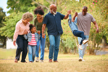 Three Generation Family Walking Outdoors Laughing And Playing And Swinging Grandchildren Between Them