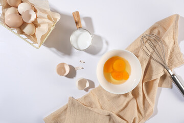 Top view of cooking layout with some ingredients on white background, a bowl of egg, a cup of milk, cream towel and a whisk. Flat lay, blank space
