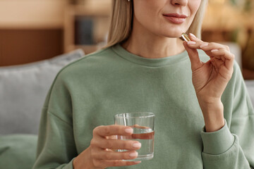 Minimal close up of adult woman taking vitamin pill and holding glass of water at home copy space