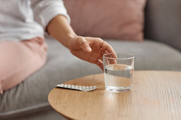 Close up of unrecognizable woman taking glass of water off wooden table with medicine pack copy space - 787060119