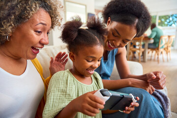 Grandmother With Mother And Granddaughter Playing With Handheld Gaming Device At Home With Family