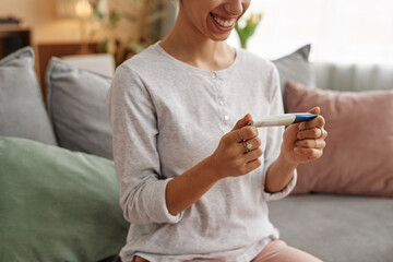 Minimal cropped shot of smiling young woman holding pregnancy test sitting on sofa at home copy space - 787061519