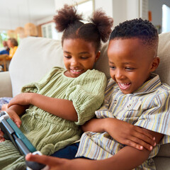 Boy And Girl Playing With Handheld Gaming Device At Home With Multi-Generation Family In Background
