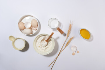 A group of dough recipe ingredients for baking cake, pastry or bread. Plenty eggs featured on round dish next to a cup of milk, a bowl of oil and flour bowl. Space for designing, top view