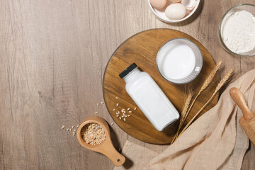 Flat lay on light wooden background with a round wooden tray contained a bottle and a glass of milk decorated by other ingredients including flour, egg, barley. Copy space, top view
