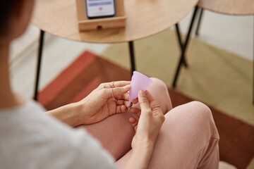 Top view closeup of female hand holding pink menstrual cup sitting on couch at home copy space