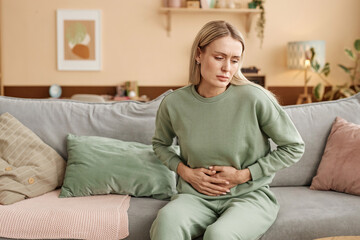 Portrait of blonde young woman holding onto stomach suffering from period cramps or pains sitting on sofa at home copy space