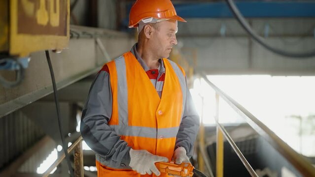 Factory worker in orange safety gear and helmet controlling cathead in workshop with tool. Industrial plant environment, machinery production