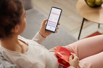 High angle closeup of young woman holding smartphone with period tracker app on screen and holding hot water bottle on belly copy space