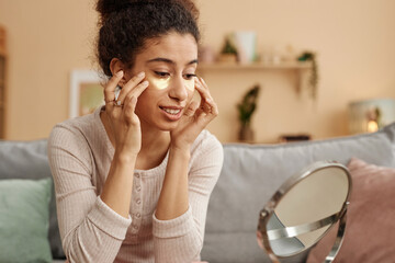 Portrait of smiling young woman applying moisturizing patches and looking in mirror at home