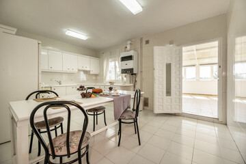 Large breakfast table with food, sweets, tablecloths and bowls in a kitchen with white lacquered furniture and integrated appliances and a metal door leading to a conservatory