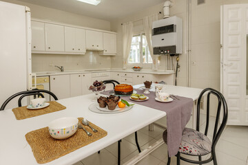 Large breakfast table with food, sweets, tablecloths and bowls in a kitchen with white lacquered furniture and integrated appliances