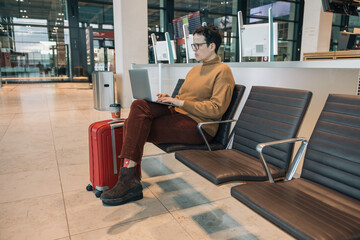 Woman with laptop sitting next to her red suitcase in airport lounge