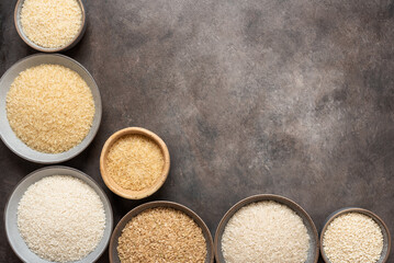 Various dry uncooked rice in bowls on a dark brown background. Top view, corner frame.