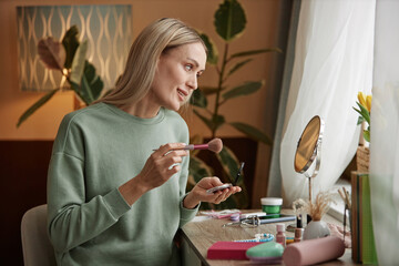 Side view portrait of blonde adult woman doing makeup at home sitting at vanity table by window copy space