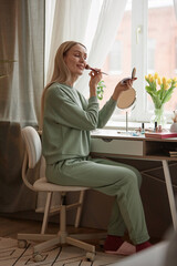 Vertical full length portrait of smiling adult woman doing makeup at home sitting at vanity table by window