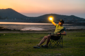 Casual man in a chair holding a lit lantern by a peaceful lake during twilight, creating a relaxing...