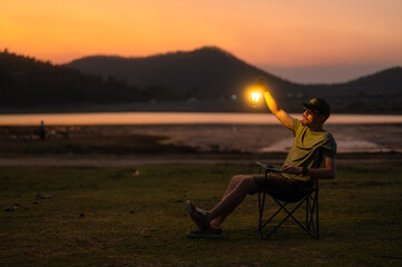 Casual man in a chair holding a lit lantern by a peaceful lake during twilight, creating a relaxing mood.