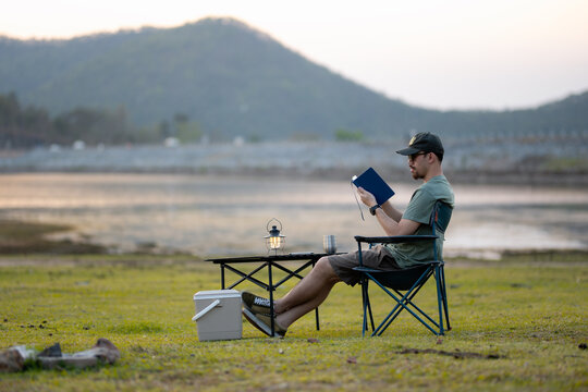 Man seated in a folding chair writing in a notebook with camping gear, enjoying the tranquility of a lakeside at dusk.