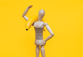 A wooden mannequin of a man holds a banana in his hands on a yellow background. The concept of healthy nutrition, fruit diet and high-calorie foods.