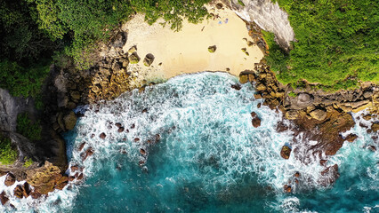 Ocean waves crash against rocks covered with greenery, forming white foam. Top view from a drone.