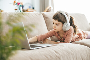 A charming little girl lies on a beige sofa, wearing headphones and watching a laptop screen....