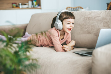 A laughing little girl with headphones watching a laptop screen, lying on a couch. Concept:...