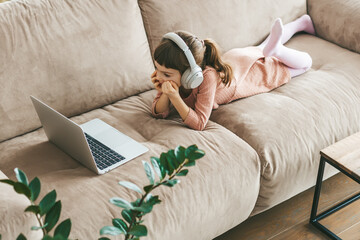 A little girl wearing headphones while watching a laptop screen, lying comfortably on a couch....