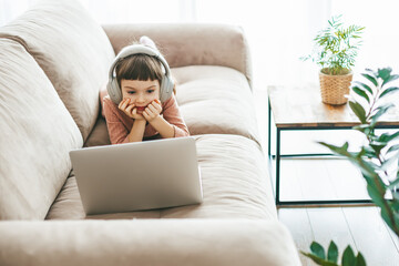 With headphones in place, a sweet little girl relaxing on a beige sofa, engrossed in her laptop...