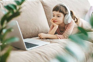A charming 5-6-year-old girl lying on a couch, wearing headphones and watching a laptop screen. Concept: technology-infused relaxation, online education, computer entertainment