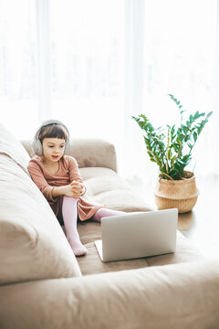 A sweet 5-6-year-old girl wearing headphones while watching a laptop, sitting on a sofa. Concept: technology-infused relaxation, online education, technological entertainment