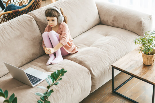 A charming little girl sitting on a sofa, wearing headphones as she watching a laptop. Concept: technology-infused relaxation, online education, technological entertainment